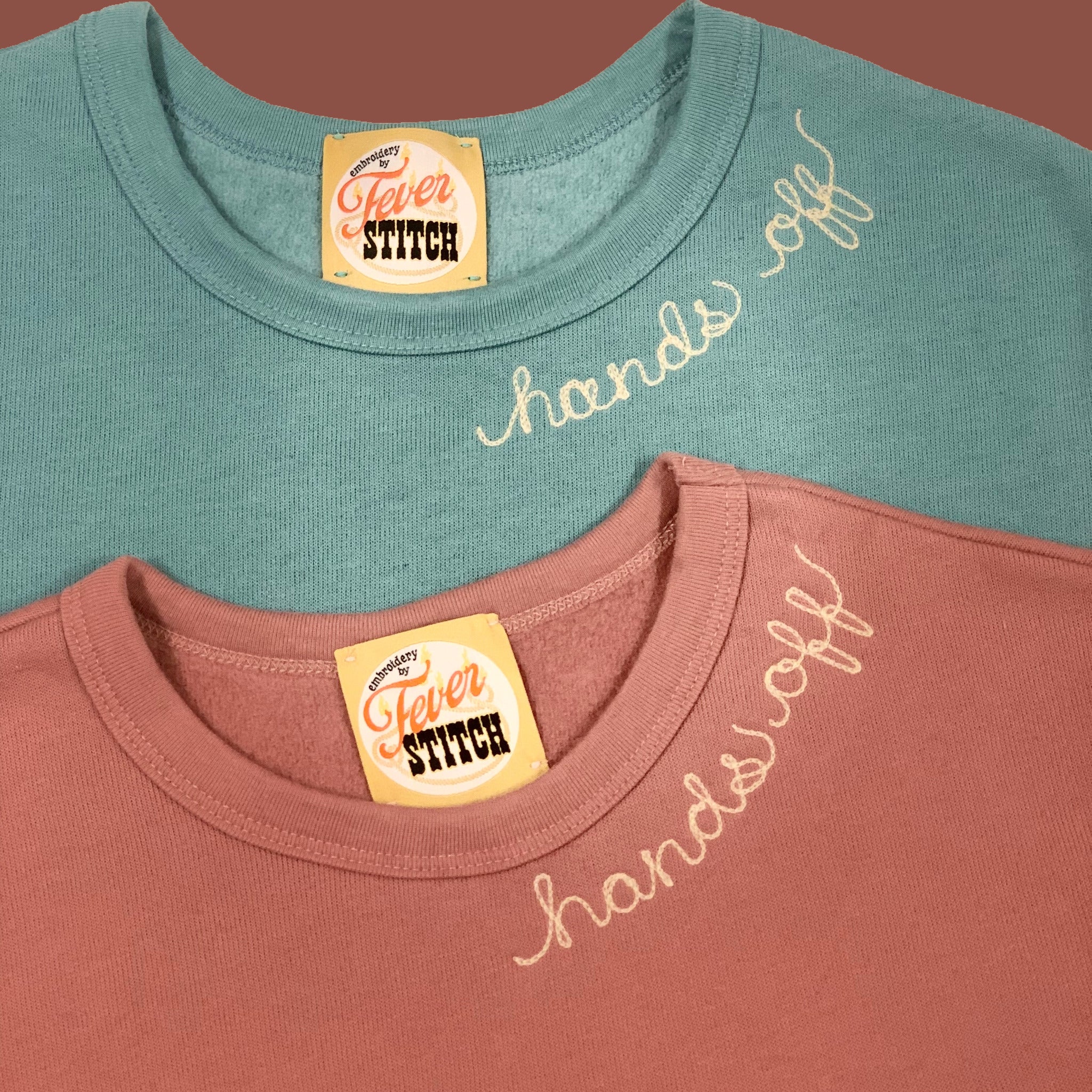 A mauve and a lagoon blue sweatshirt with the words, hands off chainstitched